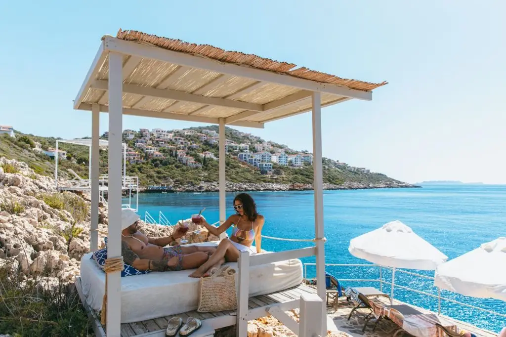 Two women enjoying a sunny beach day under a canopy, overlooking a scenic coastal town.
